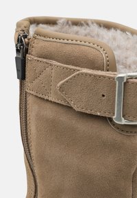 Braune Wildlederstiefel mit einem Fleece-Futter, Reißverschluss und einem Metallhardware-Riemen. Glatte Textur mit Nähakzenten.