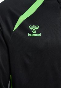 Schwarze Sportjacke mit einem grünen Streifen auf der Schulter und einem grünen hummel-Logo auf der Brust.