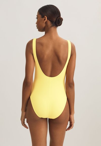 Γυναίκα με μαλλιά σε κότσο, που φοράει ένα κίτρινο ολόσωμο μαγιό με χαμηλή στρογγυλεμένη πλάτη, στέκεται μπροστά σε ένα απλό φωτεινό φόντο.