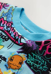 Svetlo modra majica s potiskom različnih likov Pokemon in barvnim besedilom. Rebrast kroj z okroglim izrezom in mehka tekstura tkanine.