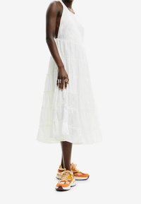 Vestido blanco de varias capas con un corpiño ajustado, que presenta un bordado intrincado. Combinado con zapatillas naranjas y blancas que muestran un diseño voluminoso.
