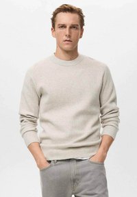 Joven con cabello castaño claro, vistiendo un suéter de canalé beige y jeans grises, de pie con las manos en los bolsillos frente a un fondo blanco liso.