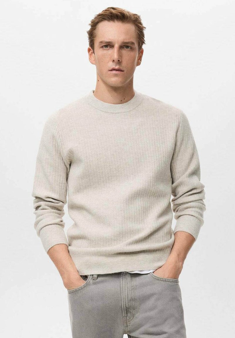 Giovane uomo con capelli castano chiaro, che indossa un maglione beige a coste e jeans grigi, in piedi con le mani nelle tasche contro uno sfondo bianco semplice.