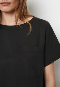 Sort kortærmet top lavet af glat stof. Har en rund halsudskæring og en enkelt påsyet lomme på venstre bryst. Minimalistisk design.