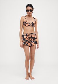 Juodas gėlėtas bikini su persikų akcentais; raštuotas viršus ir atitinkančios aukšto liemens šortai. Modelis dėvi akinius nuo saulės ir stovi basomis.