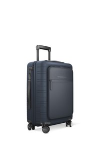 Valise rigide de couleur marine avec surface texturée, poignée rétractable, verrou intégré et quatre roues pour la mobilité. Détail de poche zippée à l'avant.