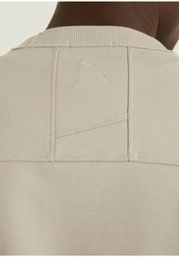 Světlé béžové tričko s texturovanou látkou, žebrovaným límcem a výrazným šitím na zádech.