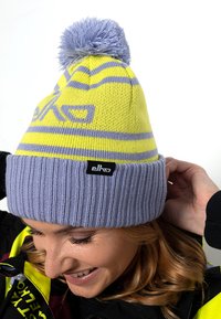 Geel en lichtpaarse beanie met een geribde rand en een pompon bovenop, met horizontale strepen en een zwart logolabel.