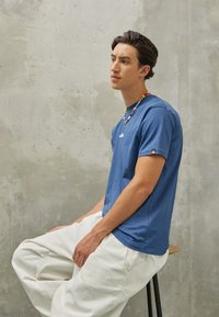 Blå bomulds T-shirt med rund hals og et lille logo, kombineret med hvide vidde bukser. En flerfarvet perlekæde tilføjer detalje.
