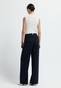 Donna con acconciatura a chignon che indossa una canotta bianca senza maniche, pantaloni larghi a righe sottili blu navy, orecchini a cerchio dorati e tacchi bianchi a punta, in piedi.