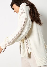 Kremowy sweter z dzianiny z teksturowymi wzorami i warkoczowymi akcentami w jasnym taupe. Posiada otwarte boki i szerokie prążkowane mankiety.