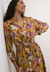 Gėlių suknele garstyčių geltonumo su violetinėmis ir rožinėmis rožėmis. Suknelė turi raukšlėtą liemenį, ilgas rankoves ir V formos iškirpimą. Tekstūruotas audinys.