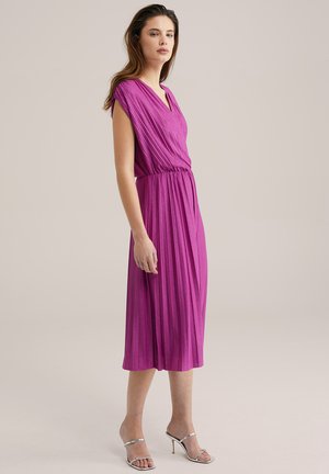 Vestido fucsia con plisados, escote en V y mangas cortas, con una cintura elástica y largo hasta la mitad de la pantorrilla, combinado con tacones plateados.
