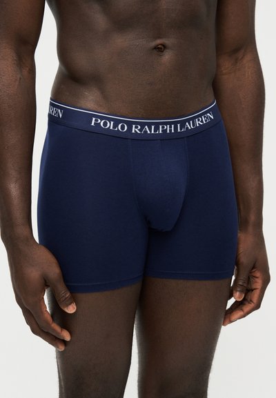 Άνδρας που φοράει σκούρο μπλε boxer Polo Ralph Lauren, τα χέρια χαλαρωμένα στο πλάι, εστίαση στον κορμό και το πάνω μέρος των μηρών.