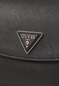 Sac à main noir texturé avec un patch logo triangulaire noir lisse et brillant présentant "GUESS USA" et un point d'interrogation en or.