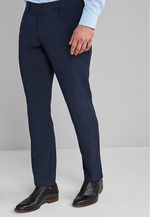 Man draagt donkere marineblauwe nette broek, zwarte leren nette schoenen en een lichtblauw overhemd met lange mouwen, staand op een effen grijze achtergrond.