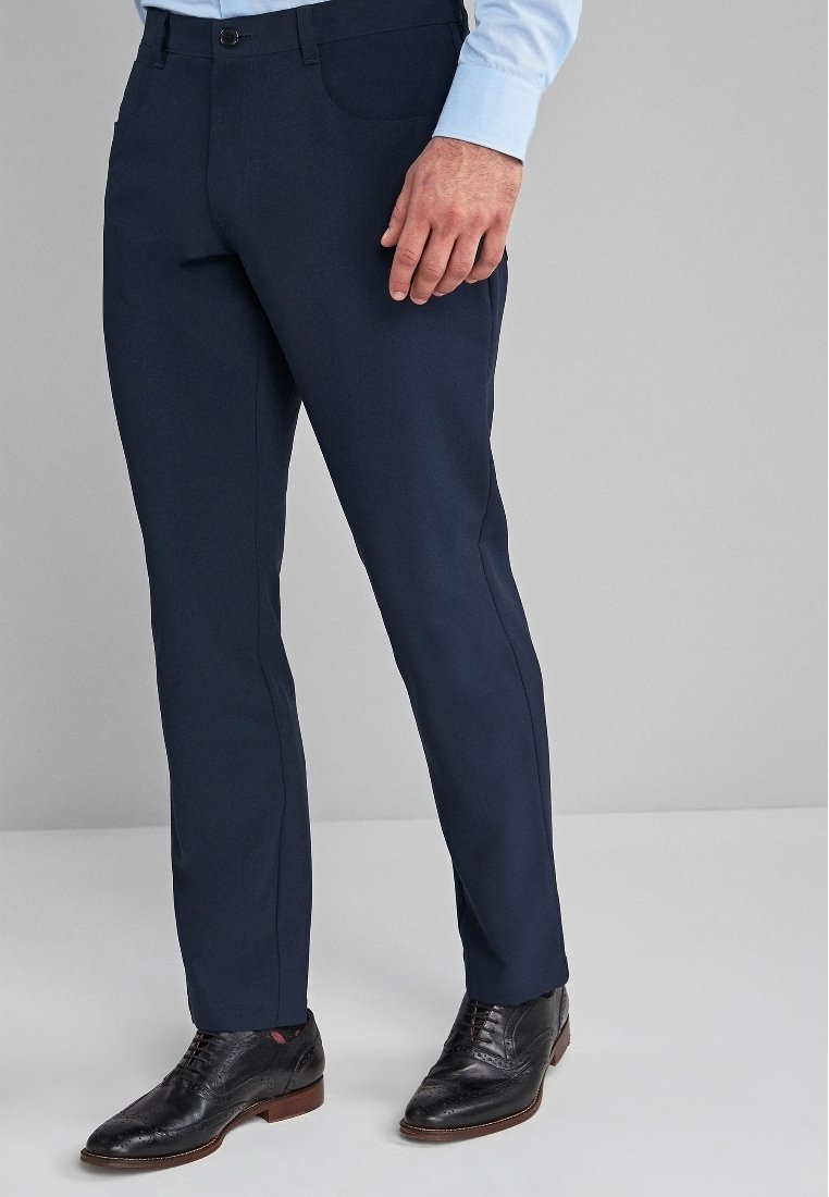 Homme portant un pantalon de costume bleu marine foncé, des chaussures de ville en cuir noir et une chemise à manches longues bleu clair, debout sur un fond gris uni.