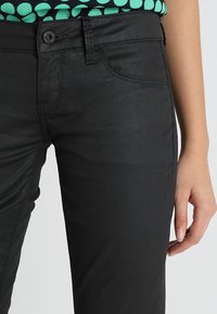 Primer plano de una persona que lleva pantalones negros con bolsillos delanteros y botón, con la mano descansando a un lado, y una blusa de patrón verde y negro.