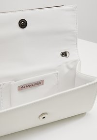 Borsa bianca con texture liscia, ferramenta argentata, chiusura a scatto e etichetta interna con la scritta "ANNA FIELD." Include una tasca per carte.