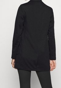 Długi czarny płaszcz o gładkiej teksturze, dopasowanym kroju i wyrazistych klapach. Design obejmuje boczne kieszenie oraz średniej długości krój.