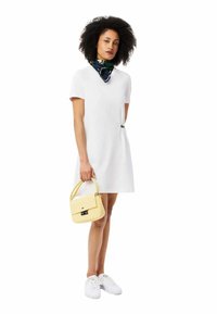 Weißes Kurzarmkleid mit lockerer Passform, das einen grünen Akzent hat. Das Modell hält eine kleine gelbe Handtasche mit einem Verschluss und trägt weiße Sneakers.