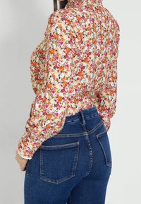 Bloes met bloemenpatroon op een witte achtergrond, met oranje, roze en groene bloemen. Gecombineerd met aansluitende donkerblauwe jeans met achterzakken.