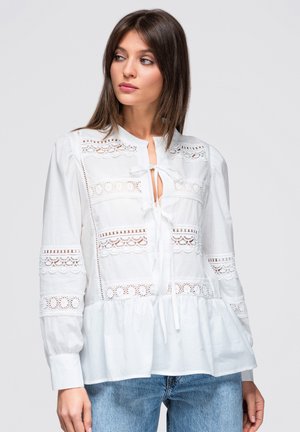 Femme portant un chemisier blanc à manches longues avec des détails en dentelle et des liens, associé à un jean bleu, regardant sur le côté sur un fond uni.