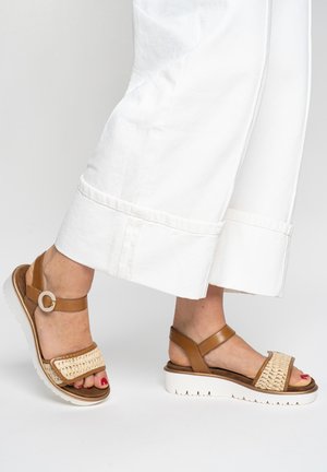 Persona con pantalones blancos de pierna ancha y sandalias de cuero marrón claro con tiras delanteras tejidas y suelas gruesas blancas.