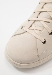 Gros plan sur la pointe d'une basket en daim beige avec des lacets beiges passés dans des œillets métalliques sur fond blanc.