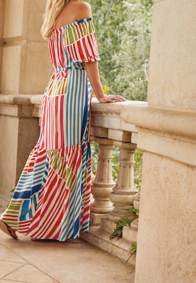 Femme portant une robe maxi rayée colorée à épaules dénudées, appuyée sur une balustrade en pierre avec des arbres en arrière-plan.