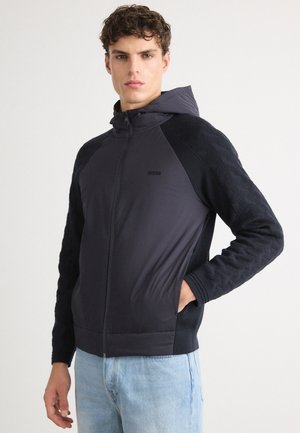 Schwarze und dunkelgraue Zip-Hoodie mit strukturierten Ärmeln, Kapuze und zwei vorderen Taschen. Verfügt über kontrastierende Materialien und minimale Markenpräsenz.