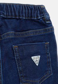 Pantaloni in denim blu scuro con vita elasticizzata, caratterizzati da una tasca posteriore e un'etichetta triangolare con il logo "GUESS" arricchita da accenti bianchi e neri.