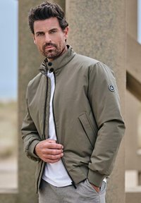 Olive green bomber jacket with a high collar, zip closure, and button accents, featuring a patch logo on the left sleeve. Worn over a white shirt.