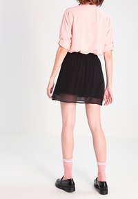 Kvinna som står vänd bort, iklädd en ljusrosa skjorta, svart kjol, rosa strumpor och svarta skor mot en vanlig vit bakgrund.