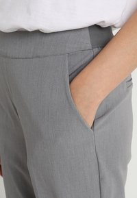 Persoon in grijze broek met een elastische tailleband, hand in de voorkant van de zak, en een wit shirt gestoken.