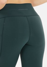 Leggings deportivos en un verde oscuro con una cintura alta, diseño sin costuras y un pequeño logo en la parte superior, fabricados con un tejido elástico y suave.