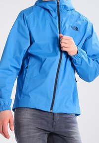 Blå vattentät jacka med huva, dragkedja fram och elastiska ärmslut. Har en svart logotyp på övre vänstra bröstet.