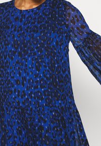 Blå klänning med leopardmönster och svarta accenter, genomskinliga långa ärmar och utsvängd kjol. Texturerad tyg med paljetter.