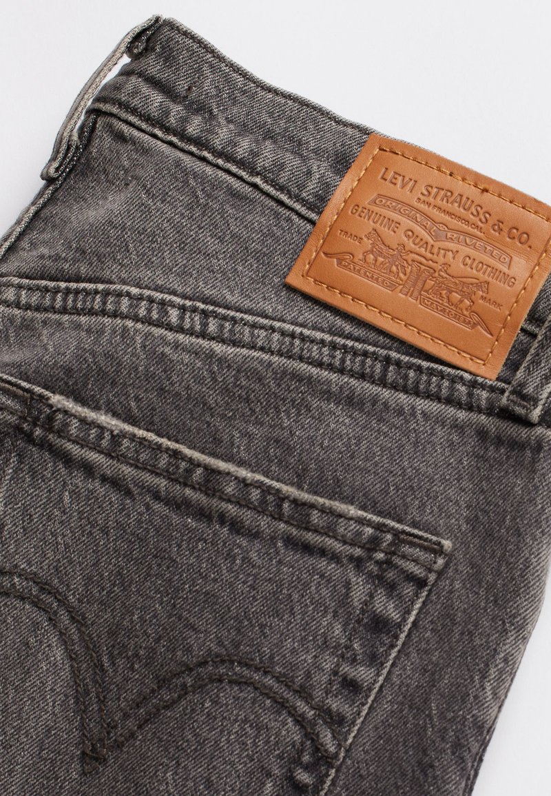 Jean en denim noir avec une étiquette en cuir présentant des détails en relief, design à cinq poches et coutures visibles sur les coutures et les poches.