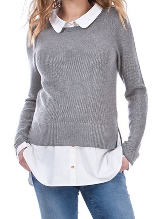 Femme portant un pull gris en maille par-dessus une chemise blanche boutonnée avec col, associée à un jean bleu.