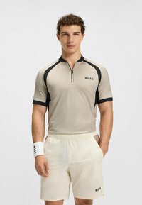 Beige kortærmet skjorte med sorte detaljer og halv-zip krave, parret med lysebeige shorts. Hvid håndledsrem med sort logo detalje.