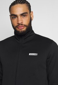 Črna jakna na zadrgo iz gladkega materiala, ki ima visok ovratnik, slim fit kroj in bel logotip na levi strani prsnega koša.