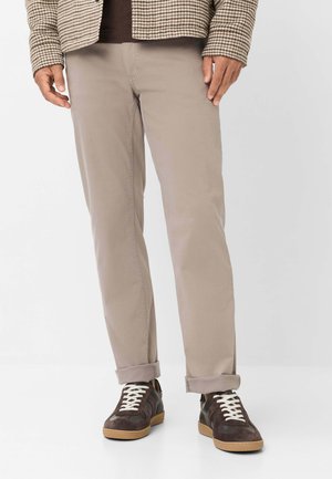 Mann mit beige-farbenen Hosen mit aufgekrempelten Säumen, braunen und weißen Sneakern sowie einer braunen karierten Jacke steht auf einem weißen Boden.