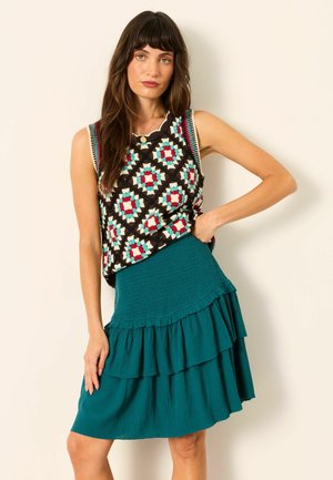 Frau mit langen braunen Haaren, die ein ärmelloses, mehrfarbig gemustertes Oberteil und einen schichtungslangen Rock in Türkis trägt, steht vor hellem Hintergrund.