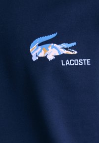 Tamsiai mėlynas medvilnės audinys su spalvinga krokodilo grafika mėlynos, baltos ir oranžinės spalvų derinyje, o žemiau matyti baltas užrašas "LACOSTE".