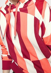 Sudadera con capucha colorida que presenta amplias rayas verticales en rojo y coral, fabricada con un tejido suave y con puños y dobladillo acanalados en contraste.