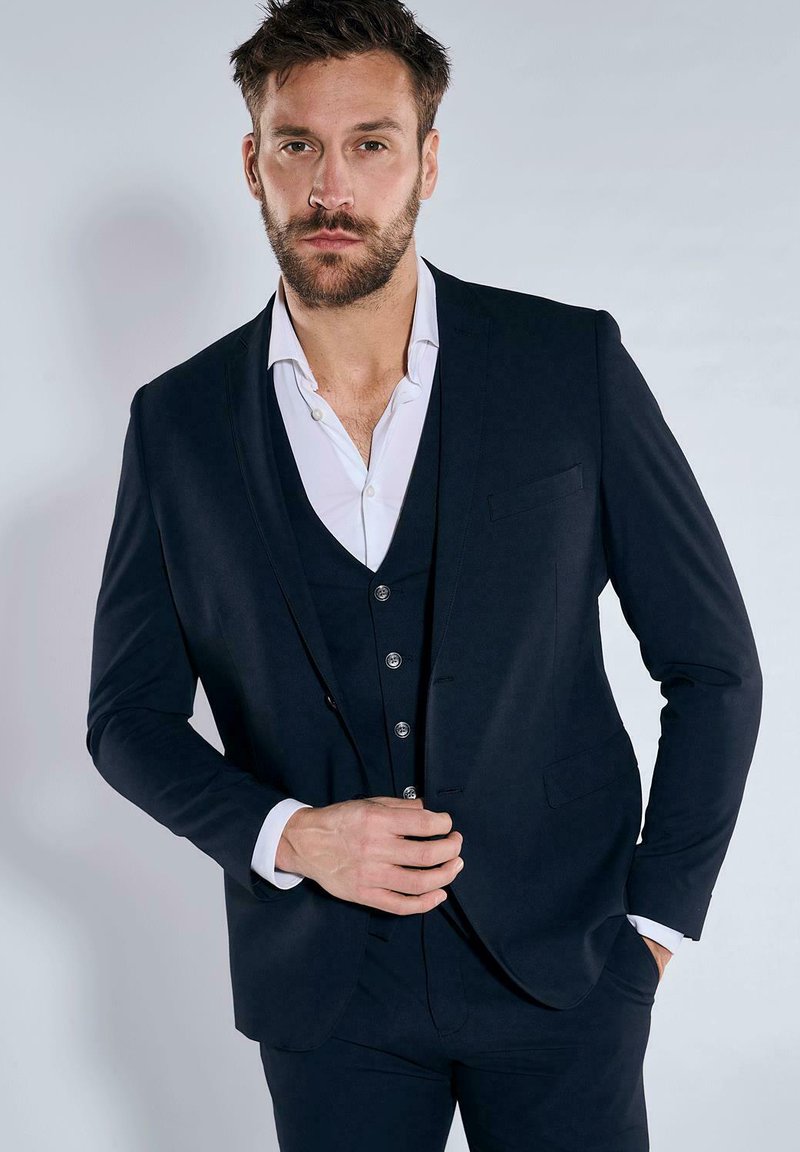 Marineblauer Anzug mit schlankem Schnitt, ausgestattet mit einem Reverskragen, einreihiger Knopfleiste und strukturiertem Stoff. Kombiniert mit einem weißen Hemd.