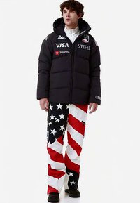 Giacca a vento nera con cappuccio, con loghi. Pantaloni bianchi con strisce diagonali rosse e blu e stelle. Scarpe nere.