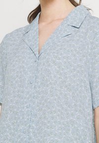 Blusa azzurra chiara con maniche corte, caratterizzata da un motivo floreale di fiori bianchi e foglie verdi, con colletto a punta e chiusura a bottoni.