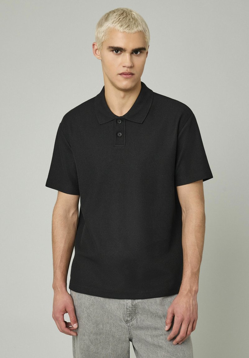 Polo noir à manches courtes avec une finition texturée, patte de boutonnage et col classique, porté avec un pantalon en tissu gris clair.